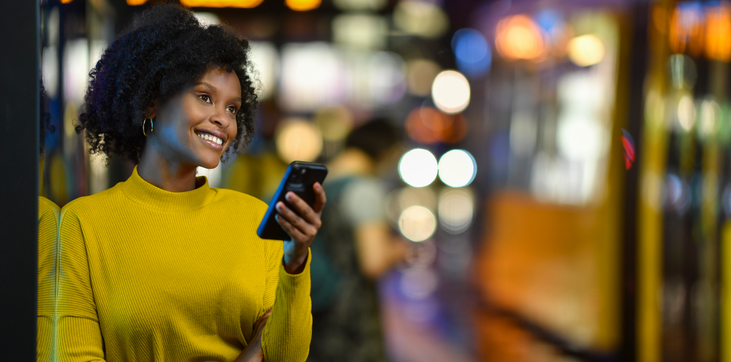 Eine junge Frau lehnt an einer Straßenbahnhaltestelle. Sie schaut dabei auf ihr Handy. Im Hintergrund ist eine Straßennbahn.