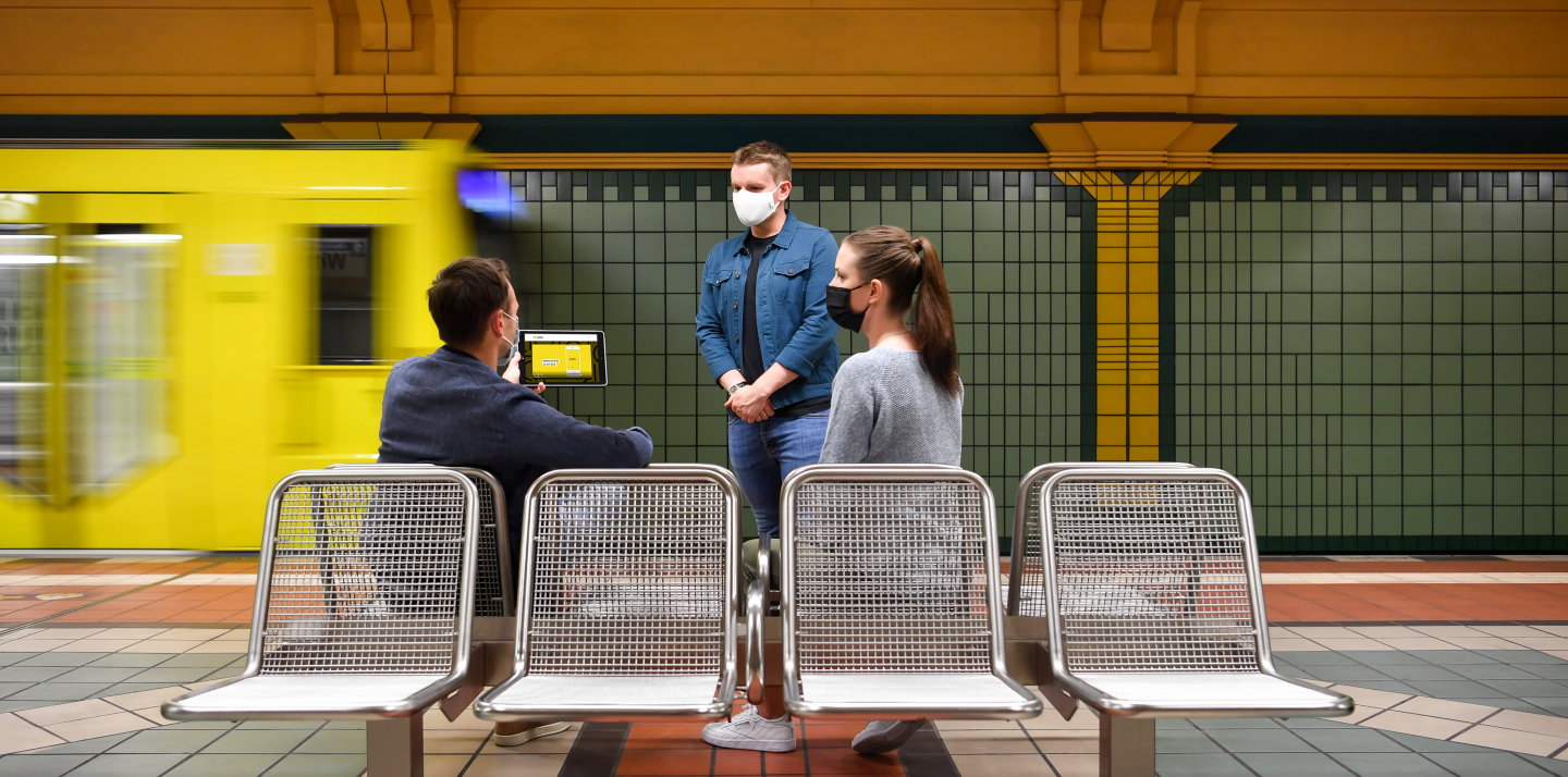 Drei Personen befinden sich in einem U-Bahnhof und schauen gemeinsam auf ein iPad. Im Hintergrund fährt eine U-Bahn aus dem Bild.