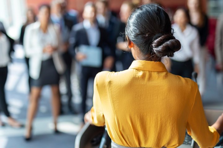 Eine Frau steht auf einem Podium. Im Hintergrund sind Frauen in Business-Kleidung zu sehen, die zuhören.